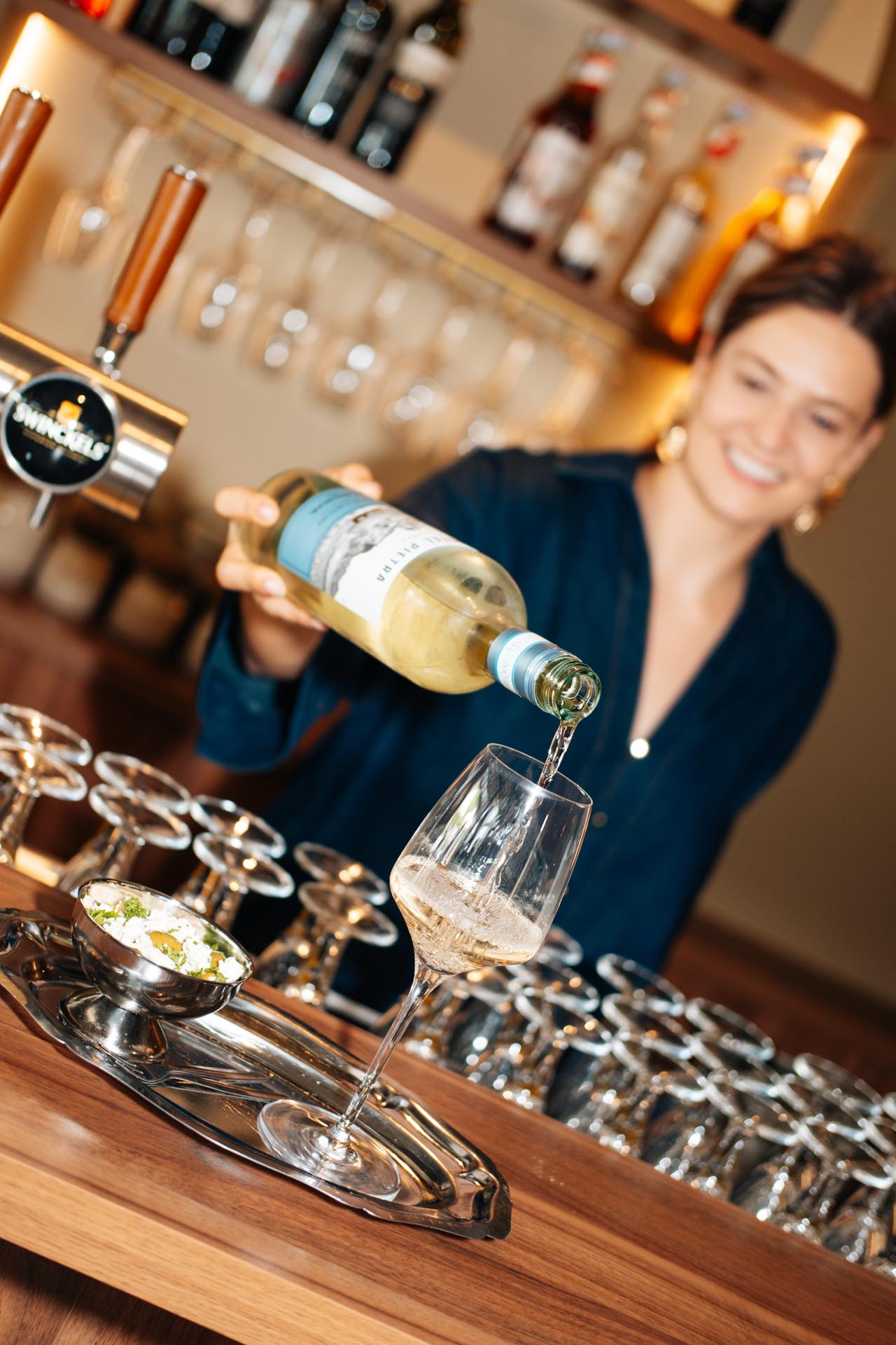Vrouw schenkt witte wijn in glas bij bar.