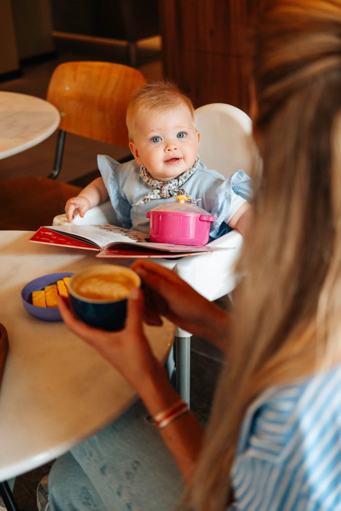 Baby in kinderstoel met speelgoed en boek op tafel.
